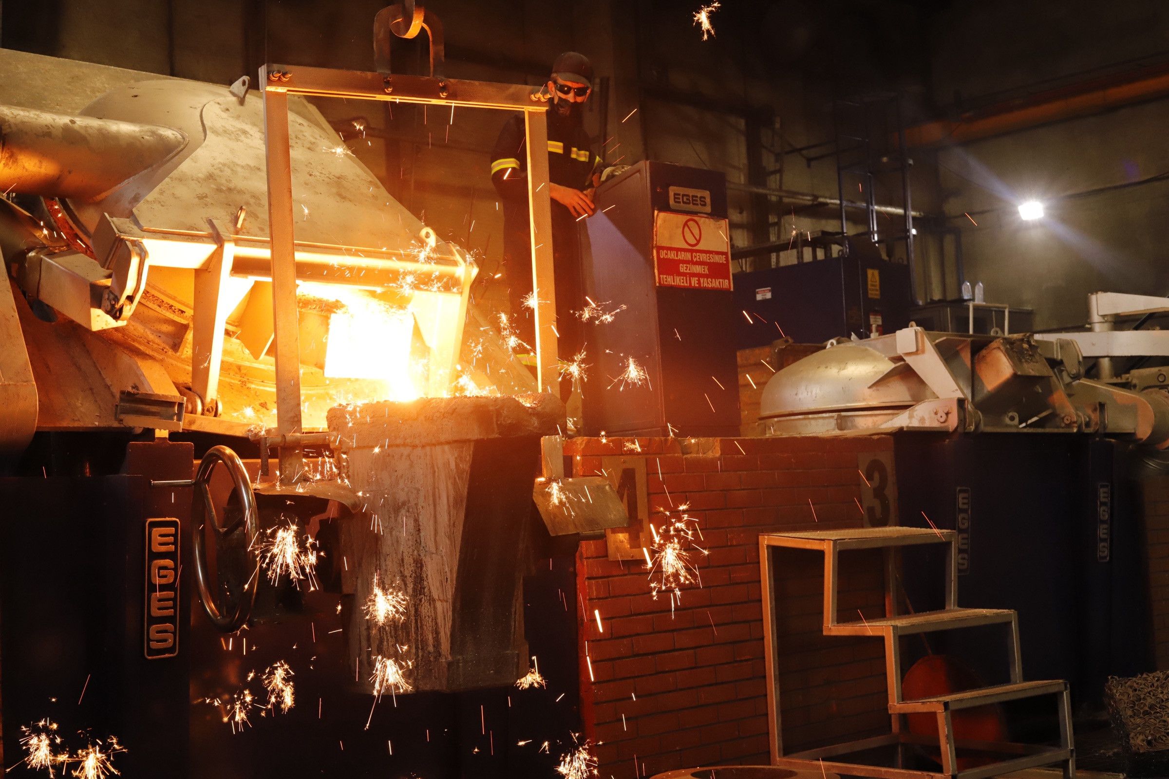 Dökümhanede indüksiyon ocağından potaya metal aktarımı; çevreye kıvılcımlar saçılıyor