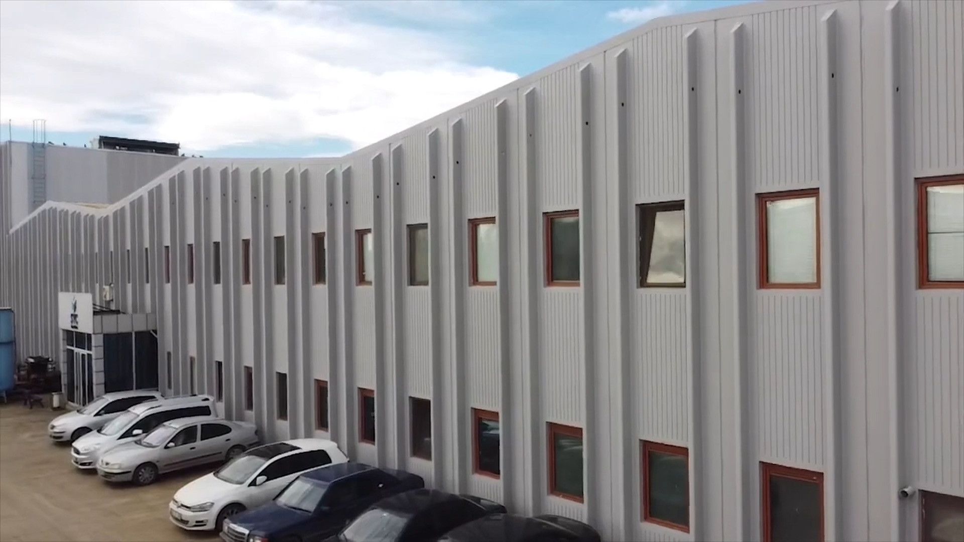 Tesisin dış cephesi: açık gri panel kaplı iki katlı bina ve önünde park etmiş araçlar